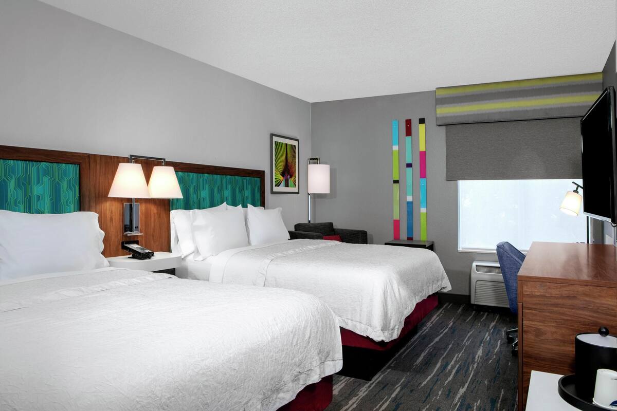 A hotel room with two beds, a desk, a chair, a TV, a lamp, a picture on the wall, and a window with a view.