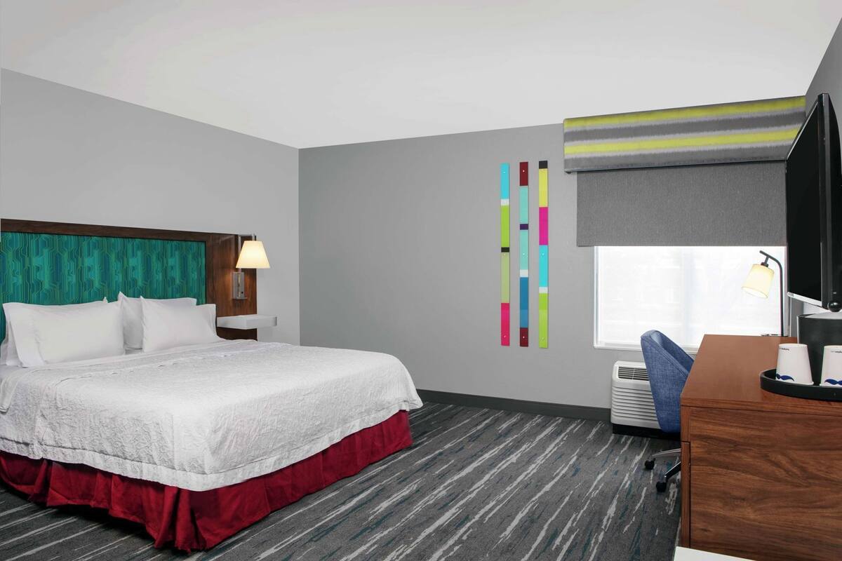 A hotel room with a bed, a desk, a chair, a window with a view, a wall with colorful vertical stripes, and a lamp.