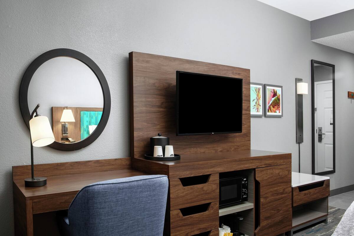 A modern hotel room with a wooden desk, a flat-screen TV, a round mirror, and a small side table with cups.