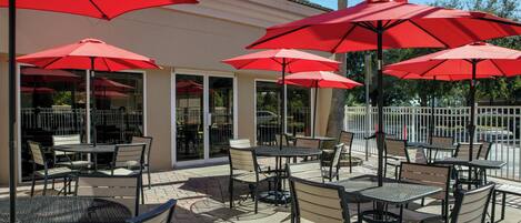 An outdoor patio with tables and chairs, red umbrellas, and a building with large windows.