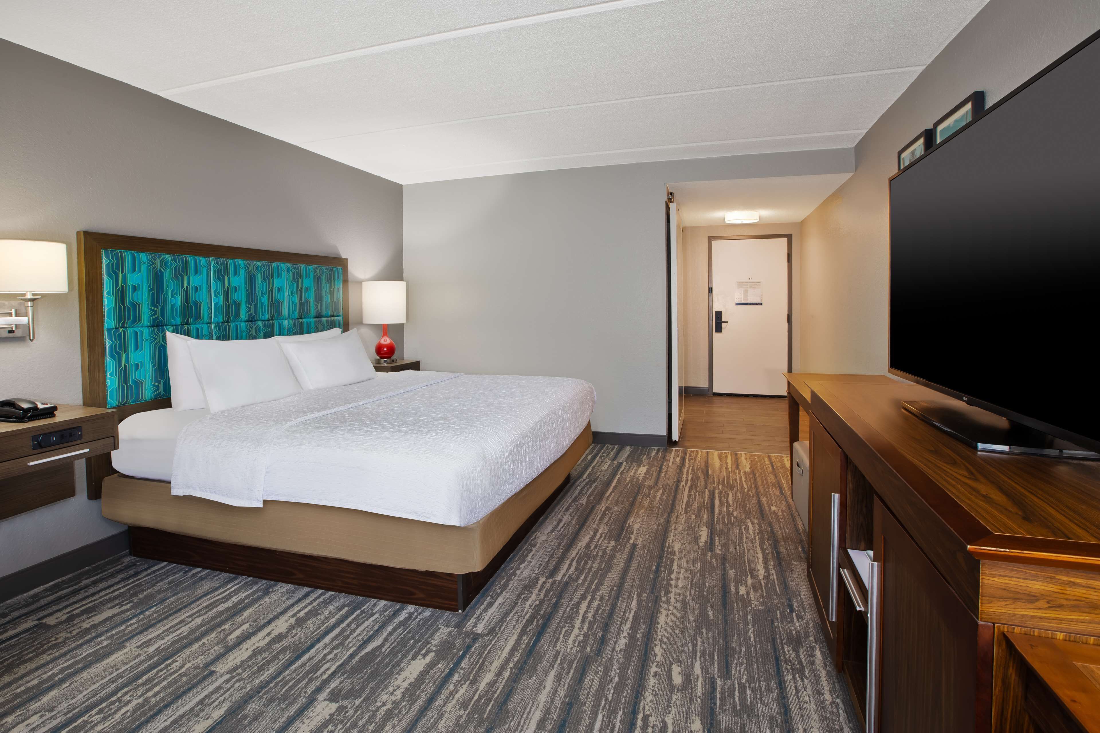 Una habitación de hotel con una cama grande, un cabecero de madera, un televisor de pantalla plana, un aparador de madera y una mesita de noche con lámpara.