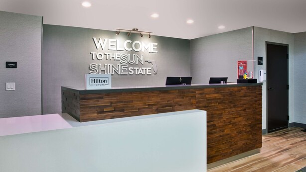 A hotel reception desk with a "Welcome to the Sun Shine State" sign, a "Hilton Honors" logo, and a modern design with a wooden accent wall. - Hampton Inn Daytona/Ormond Beach (Ormond Beach)