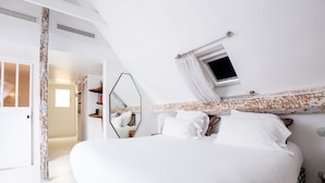 A bedroom with a large bed, a mirror, a small window with curtains, and a door leading to another room.