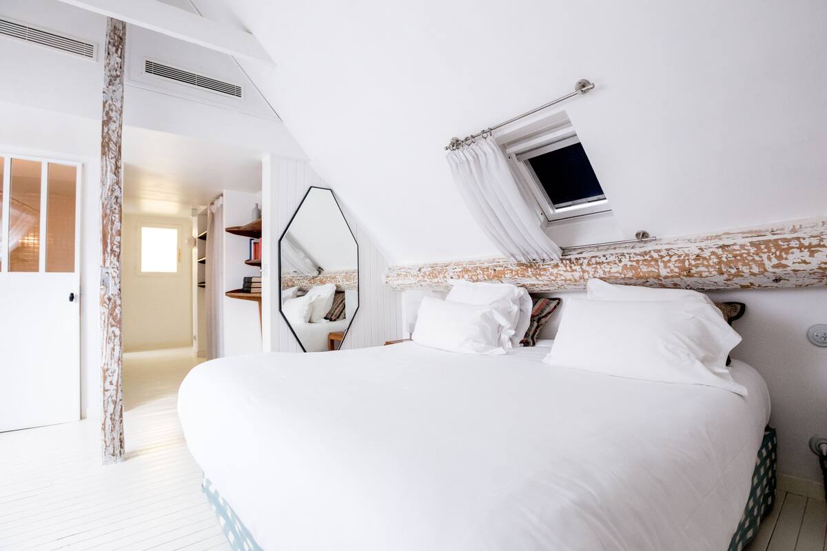A bedroom with a large bed, a mirror, a small window with curtains, and a door leading to another room.