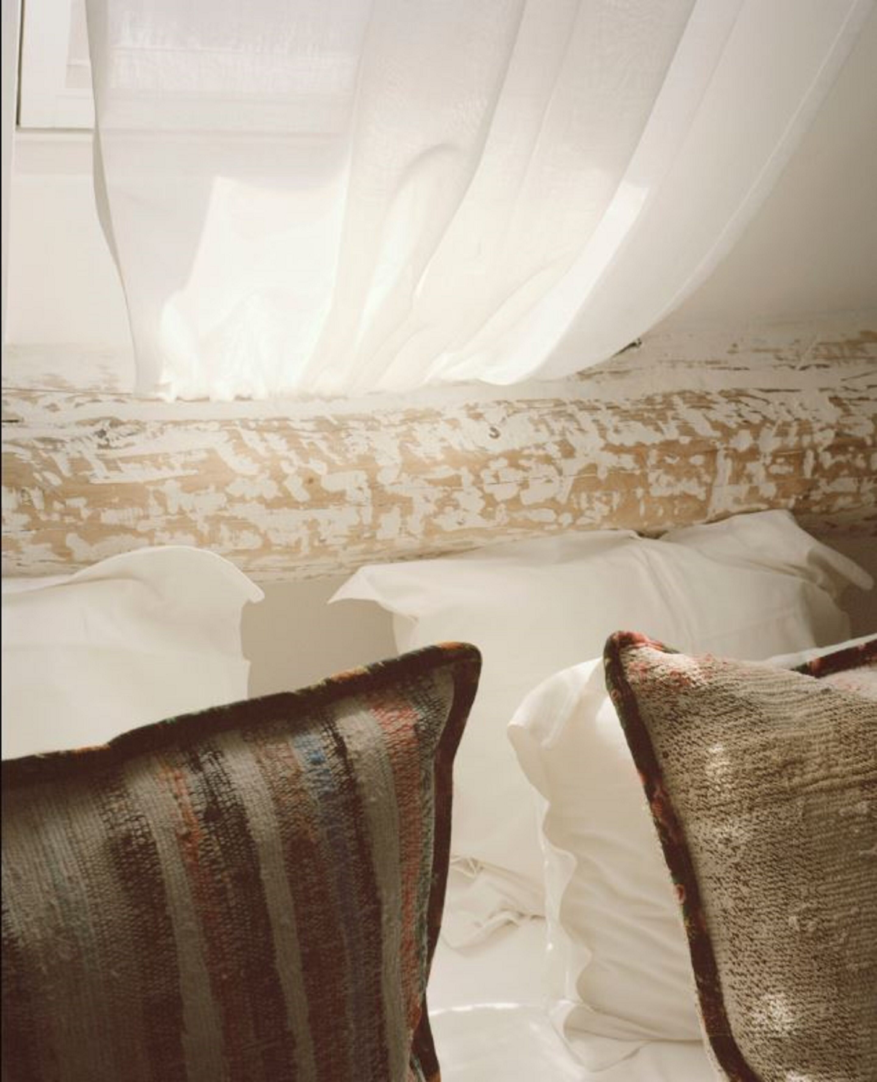 A bed with white linens and a patterned pillow, a sheer white curtain, and a textured wall.