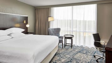 Ein Hotelzimmer mit einem großen Bett, einem Schreibtisch, einem Sessel und einer Aussicht auf die Stadt durch das Fenster.