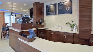 A modern reception area with a wooden counter, a coffee machine, and a menu board displaying food and beverages.