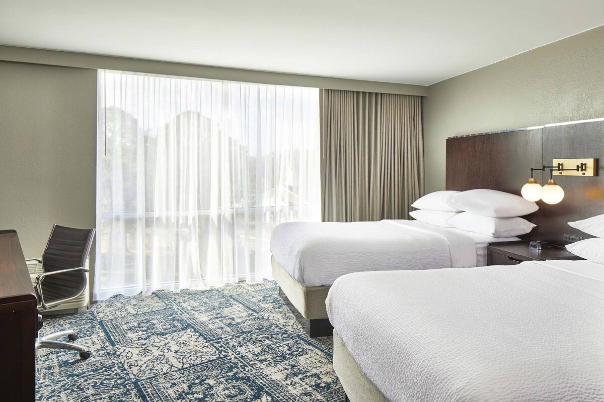 Habitación de hotel con dos camas, un escritorio, una silla, un ventanal grande con cortinas y una alfombra estampada.