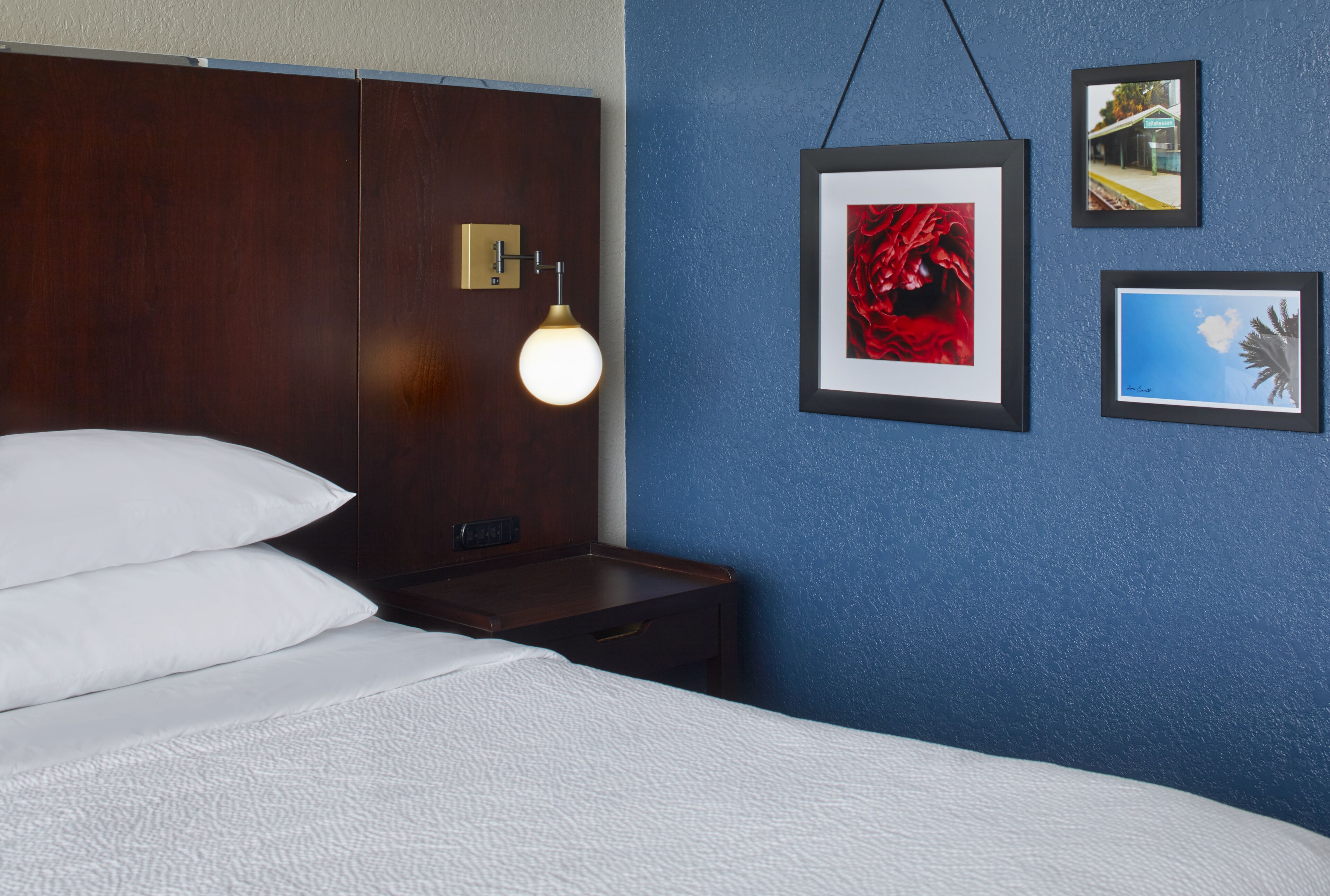 A hotel room with a bed, a nightstand, a wall-mounted lamp, and framed pictures.