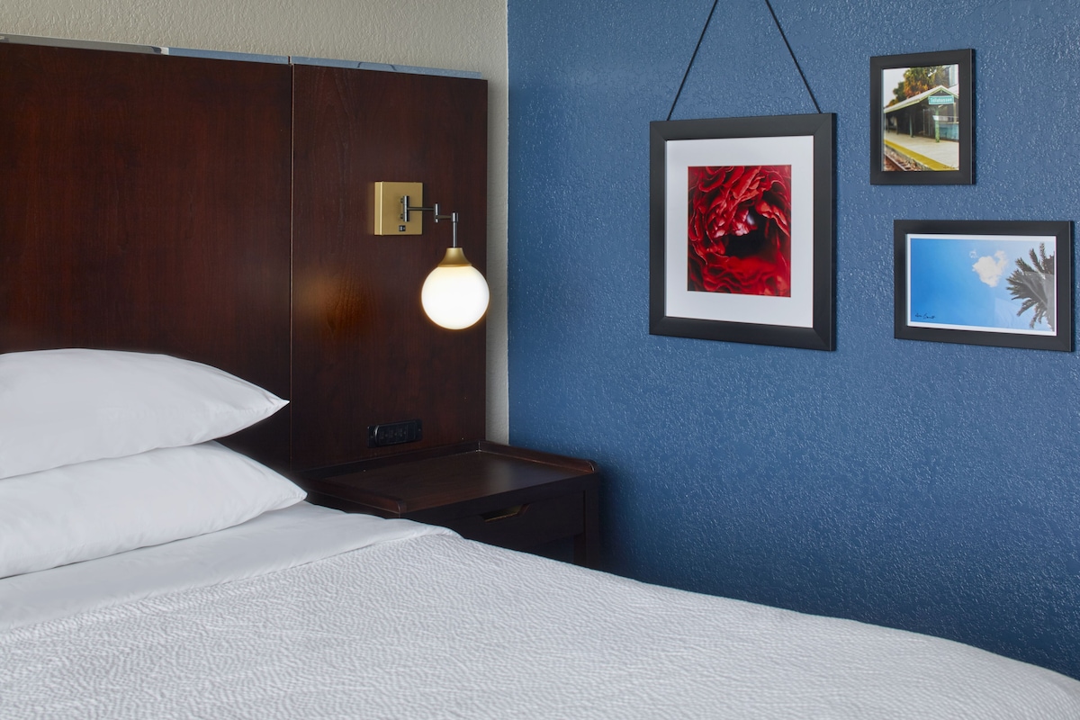 Une chambre d’hôtel avec un lit, une table de chevet, une lampe fixée au mur et des tableaux encadrés.