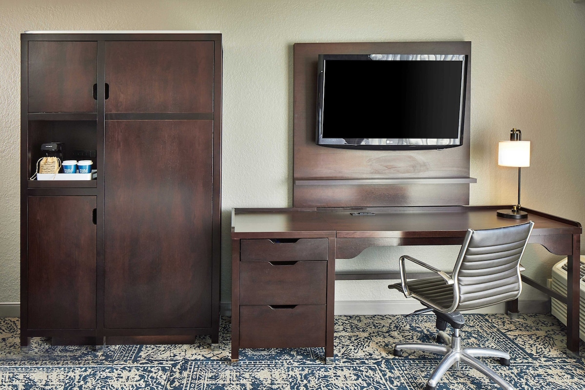 Une chambre d’hôtel avec un bureau en bois foncé, une chaise moderne, un téléviseur fixé au mur et une petite étagère sur laquelle se trouvent une machine à café et des tasses.