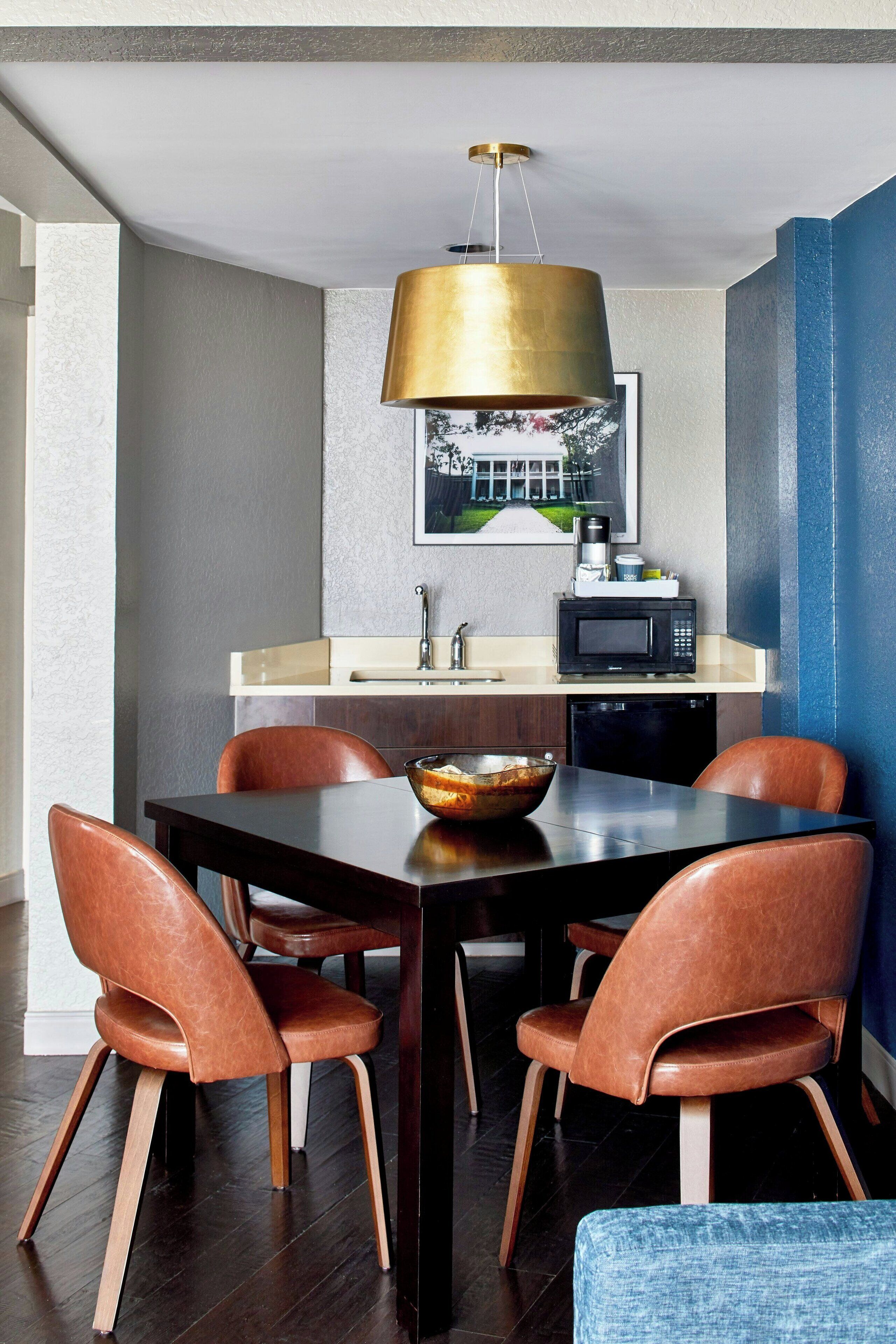 Un coin repas comprenant une table en bois foncé, quatre chaises en cuir marron, un luminaire suspendu doré, un four à micro-ondes et un évier.