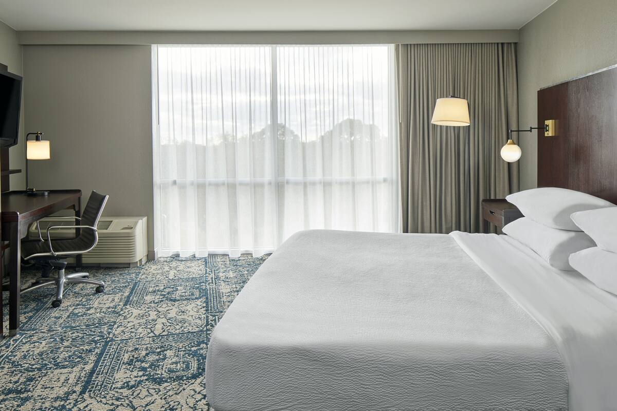 Habitación de hotel con una cama grande, un escritorio con silla, un televisor y un ventanal con cortinas.