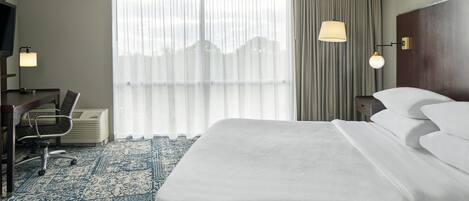 A hotel room with a large bed, a desk with a chair, a television, and a large window with curtains.