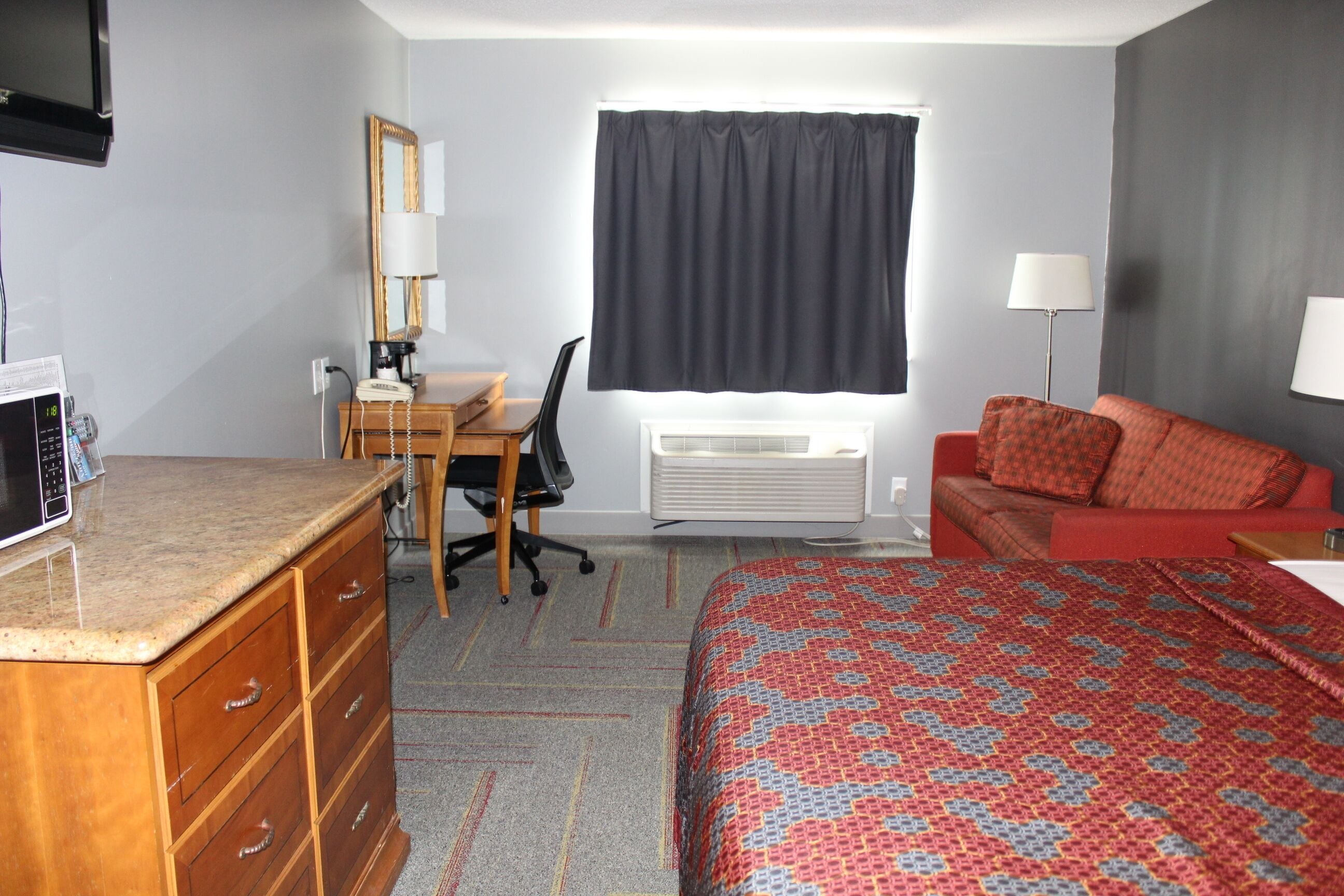 Habitación de hotel con cama, escritorio con silla, cómoda, televisión, ventana con cortinas y lámpara.