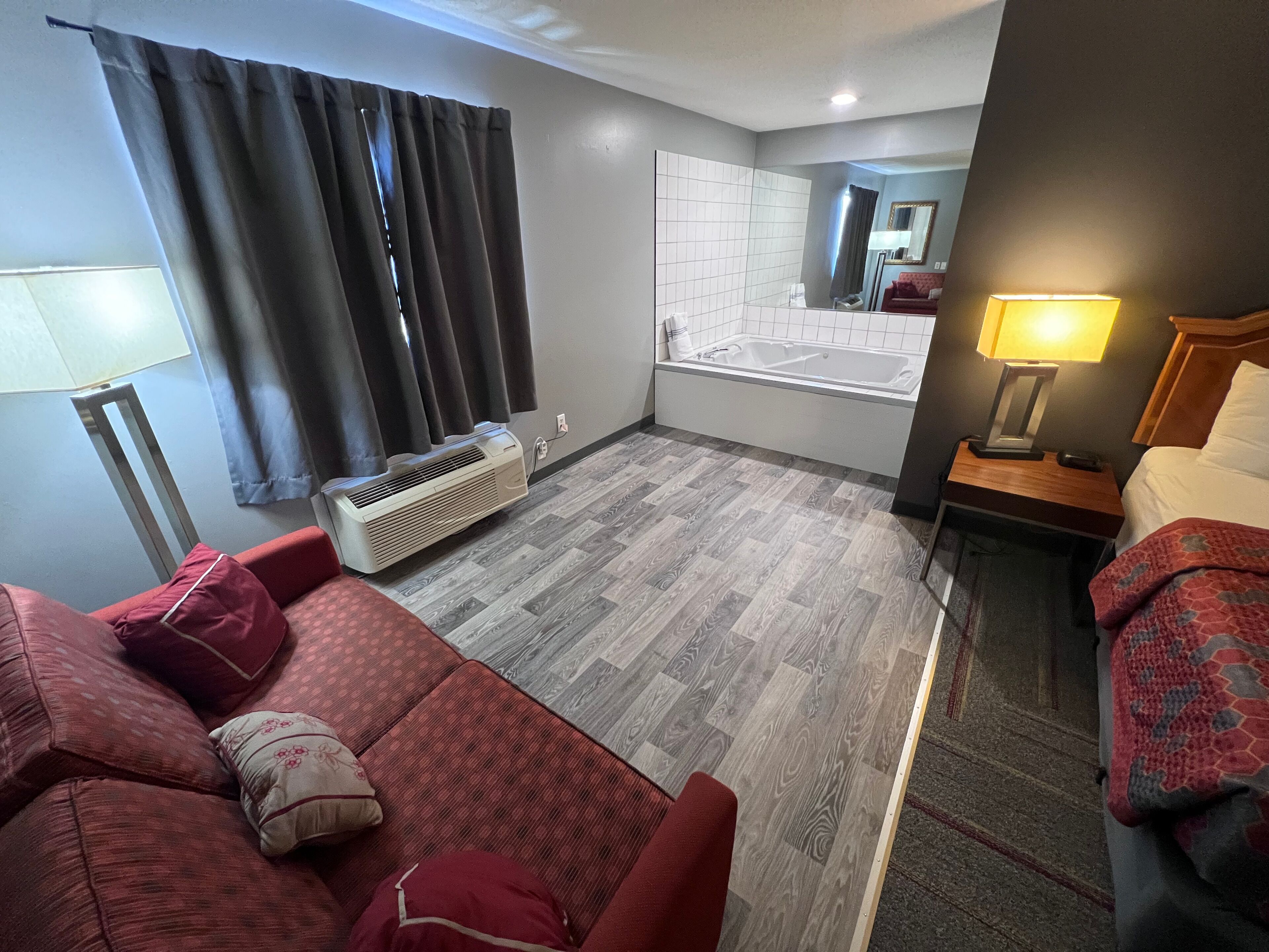 A hotel room with a red sofa, a small bedside table with a lamp, a bed with a patterned blanket, a bathroom with a bathtub, and a heater on the floor.