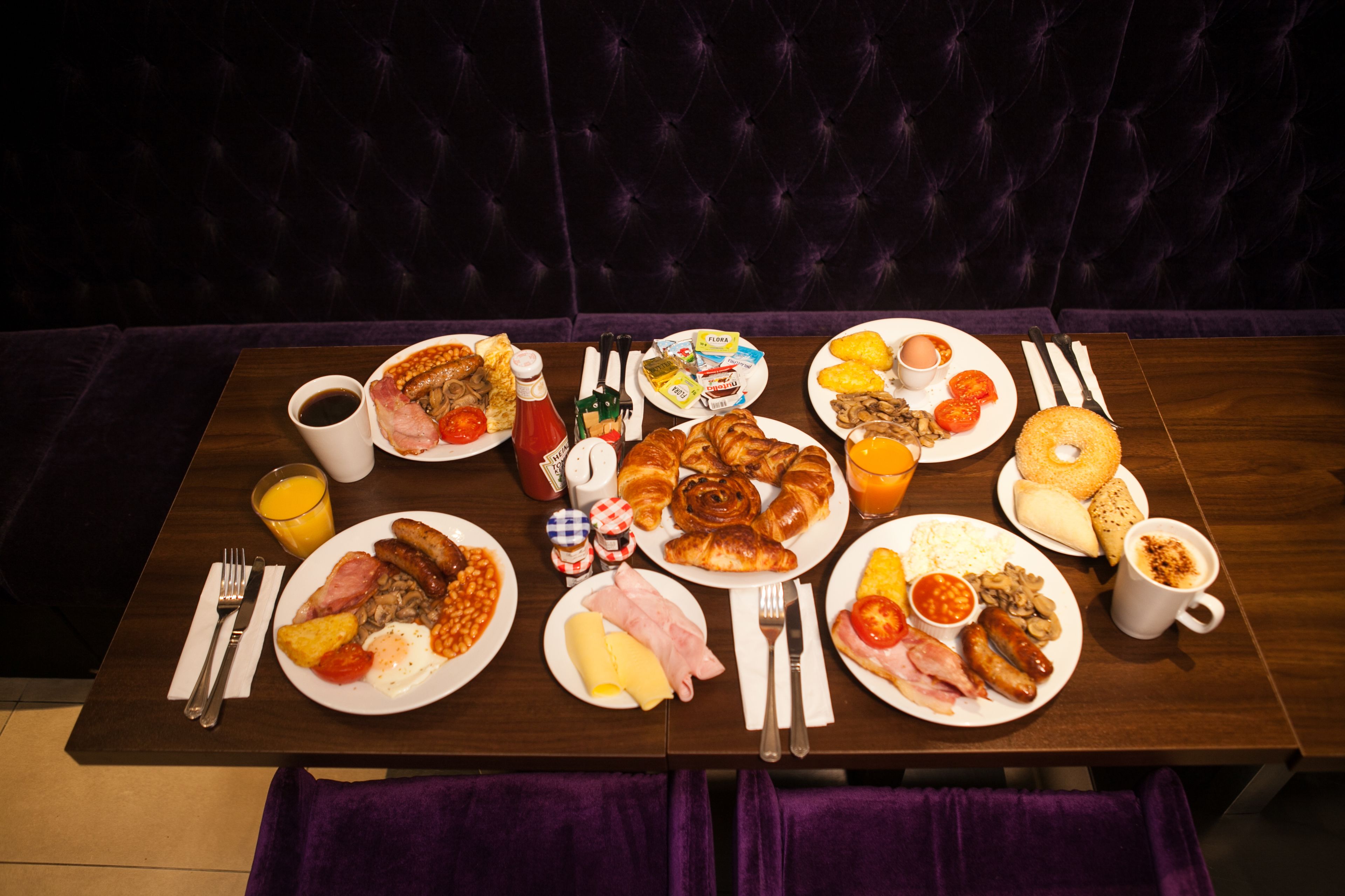 A breakfast table with various dishes, including eggs, sausages, and pastries, set on a wooden table with a purple upholstered bench in the background.