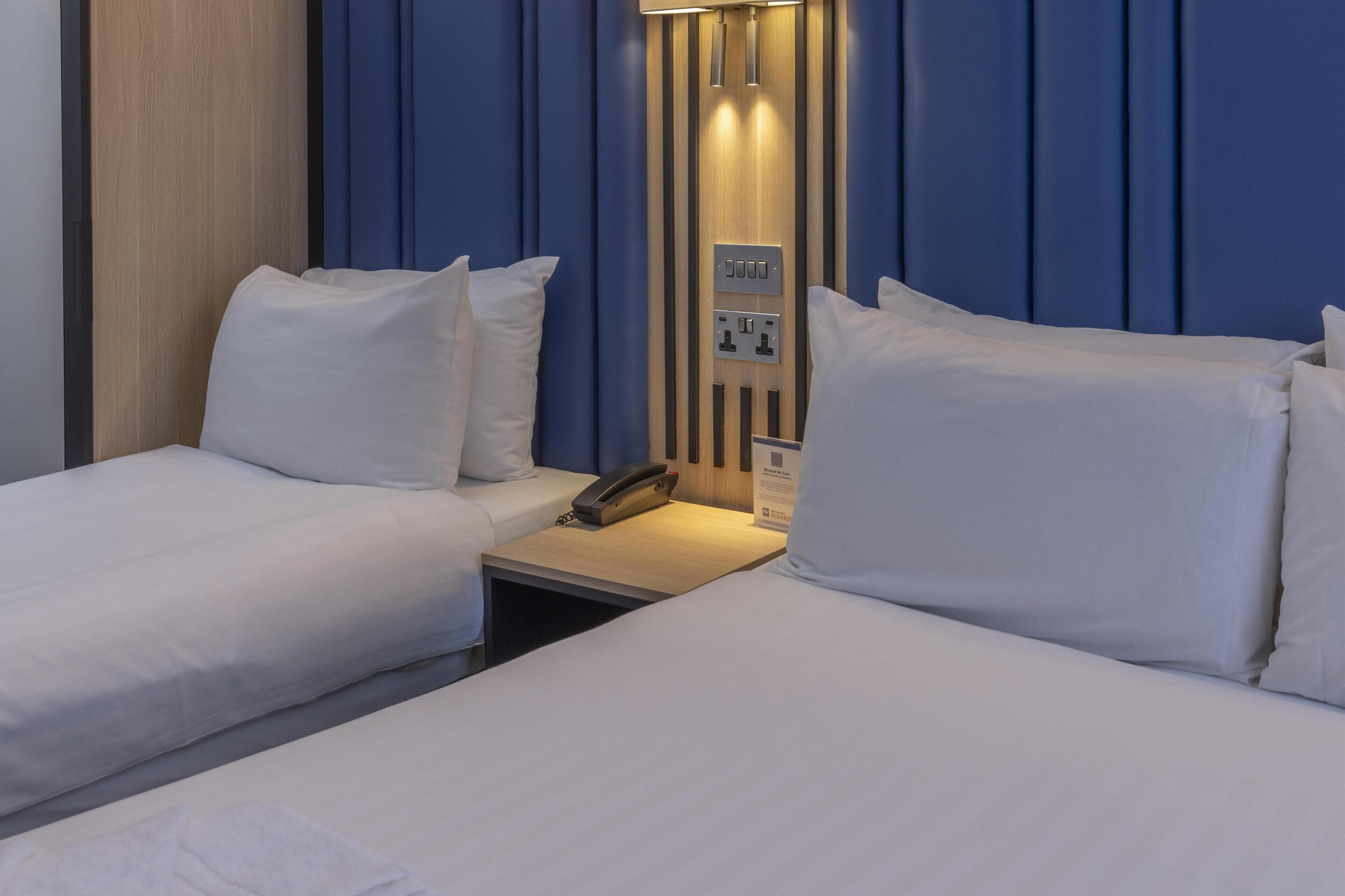 A hotel room with two beds, a bedside table with a phone, and a wall-mounted control panel.
