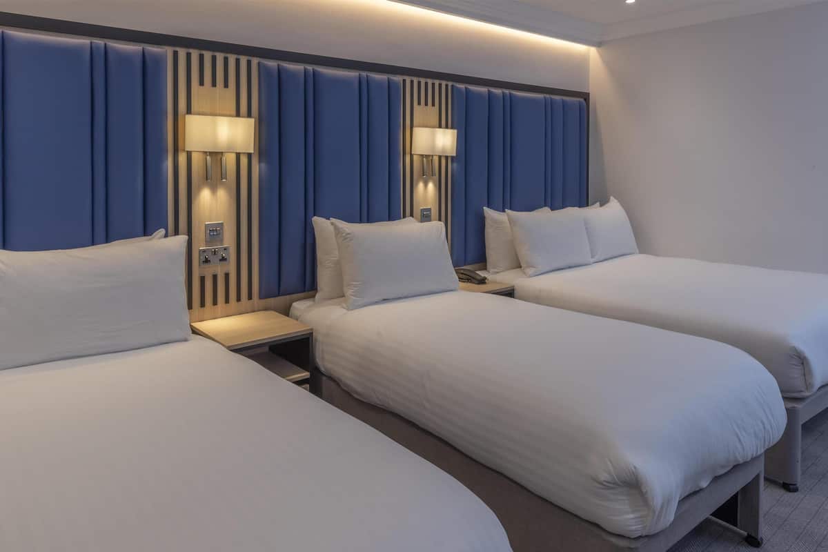 Een hotelkamer met drie bedden, een modern hoofdeinde, bedlampen en een bedieningspaneel.