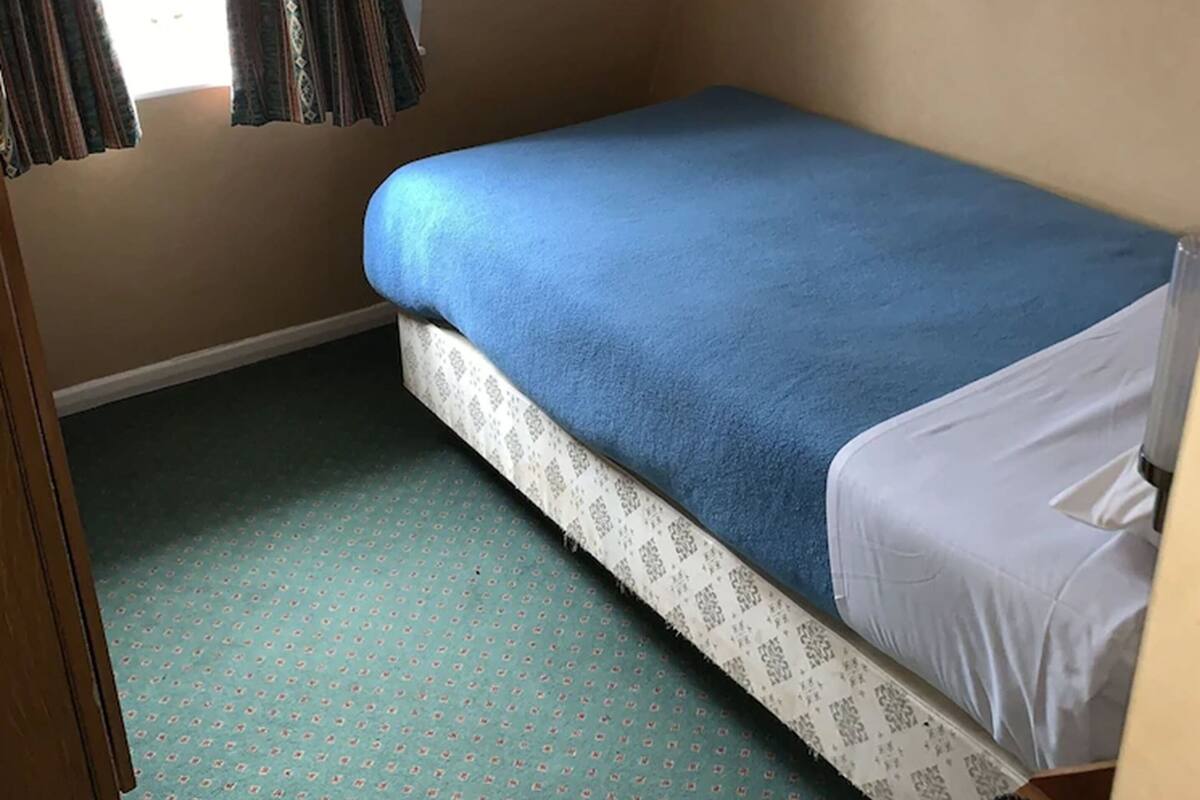 Pequeña habitación con una cama individual, colcha azul, alfombra estampada y una ventana con cortinas.