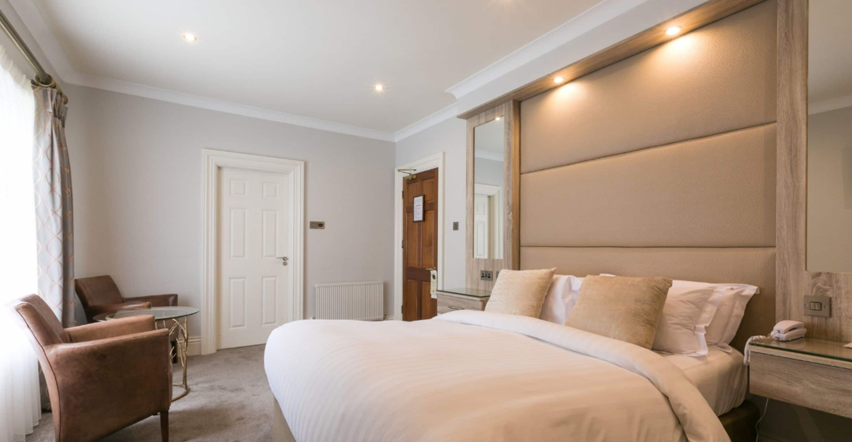 A hotel room with a large bed, a sitting area with a chair and table, a mirror, and a bedside table with a lamp.