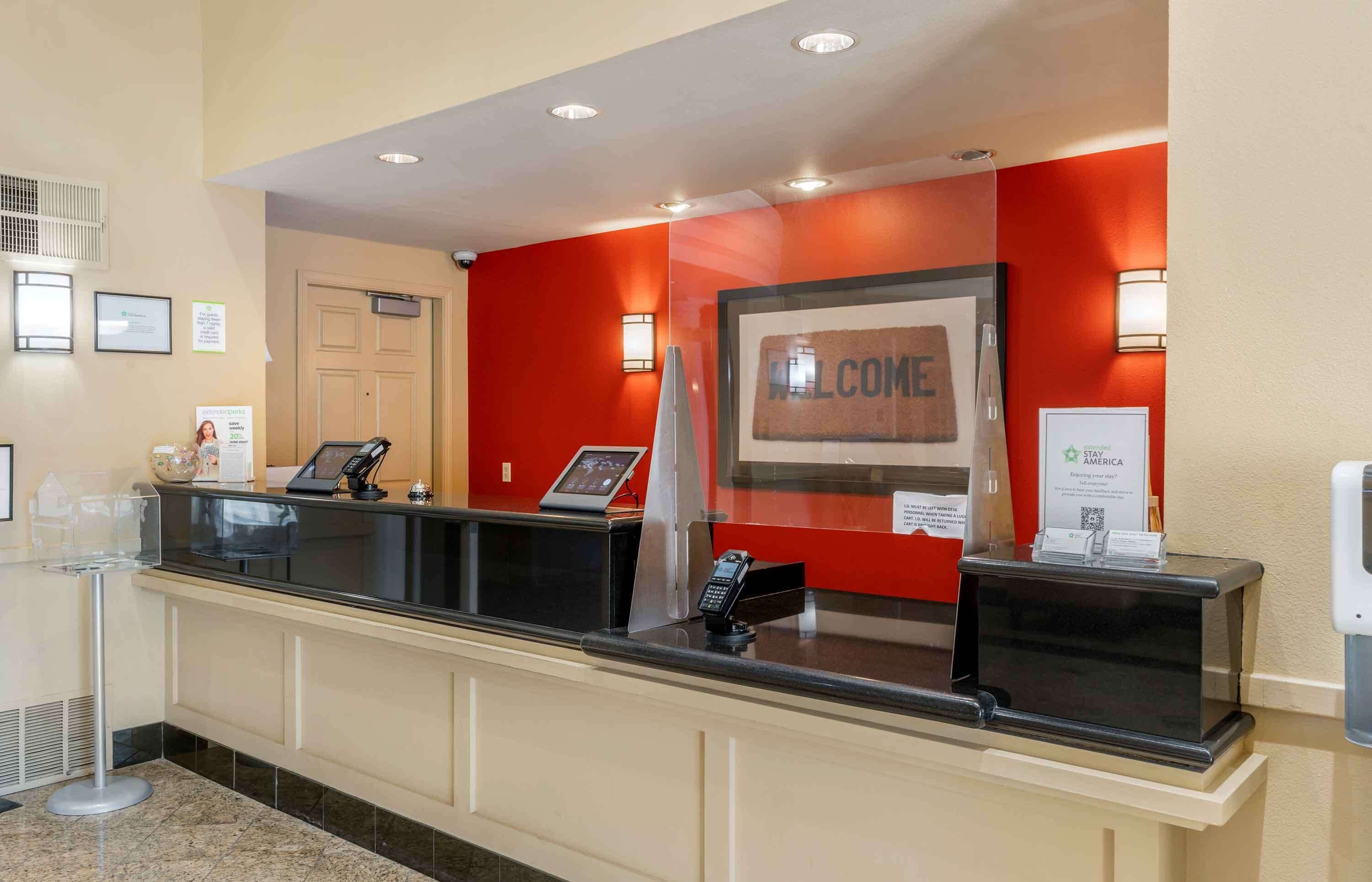 Zona de recepción de un hotel con una pared roja, un cartel de bienvenida, un mostrador de recepción y un letrero de 'Stay America'.