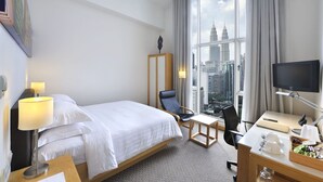 A hotel room with a bed, desk, chair, and large window with a city view.