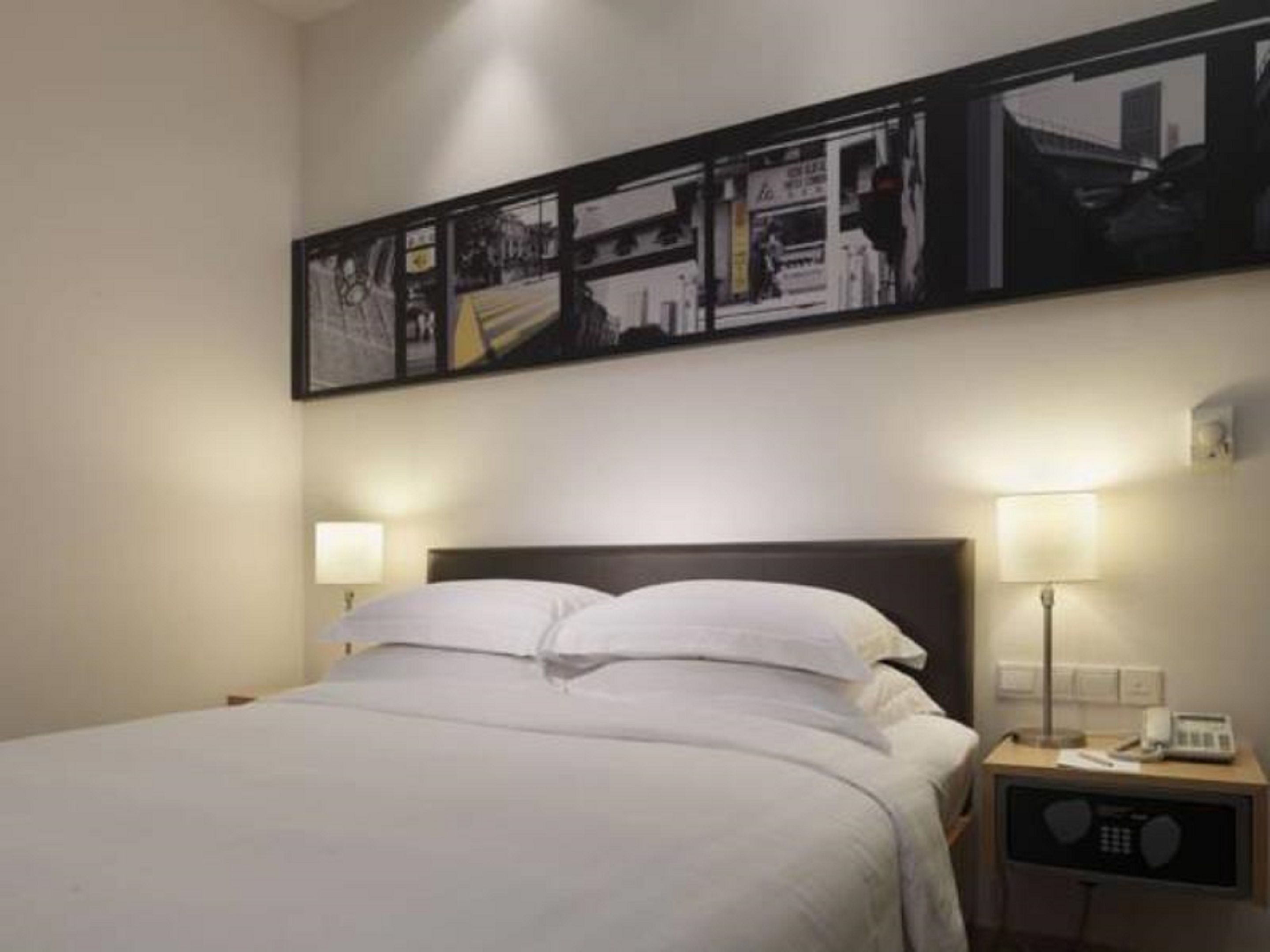 A hotel room with a bed, two bedside lamps, a nightstand with a phone, and a wall-mounted picture.