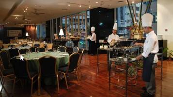 Un restaurant doté de nombreuses tables et chaises, d’une cuisine où travaillent des chefs, et de grandes fenêtres.