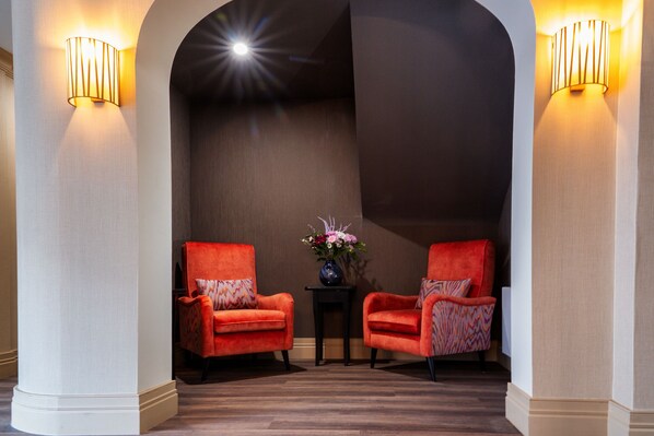 Un hall d’entrée avec deux fauteuils rouges, une petite table sur laquelle est posé un vase de fleurs et des luminaires fixés au mur.