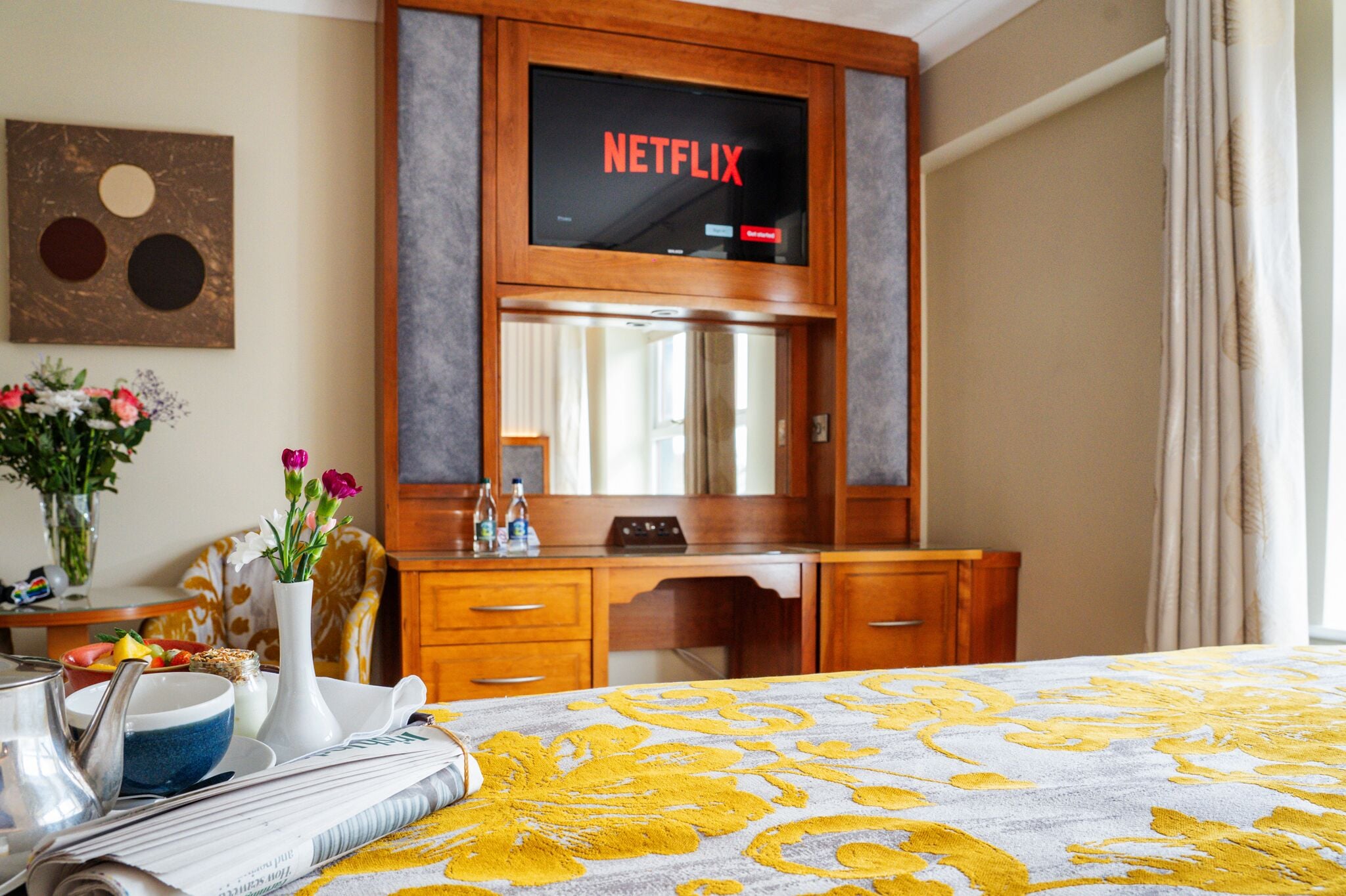 Ein Hotelzimmer mit einem Bett, einem Holztisch, einem Spiegel, einer Vase mit Blumen, einem Tablett mit Frühstücksangeboten und einem Fernseher mit dem Netflix-Logo.