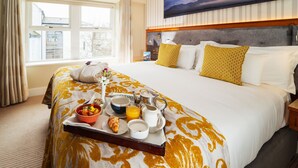 Habitación de hotel con una cama grande, un carrito de desayuno con comida y bebidas, un cuadro en la pared y una ventana con cortinas.