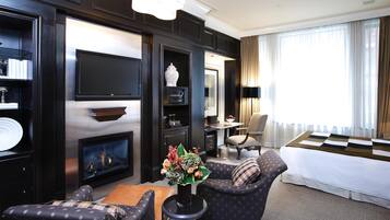 A modern hotel room with a fireplace, comfortable seating, a flat-screen TV, and a large window with curtains.
