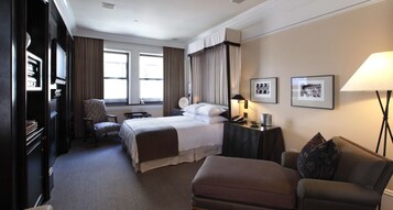 A hotel room with a large bed, a desk, a chair, a sofa, and a lamp. There are two windows with curtains, a wall-mounted TV, and framed pictures on the wall.