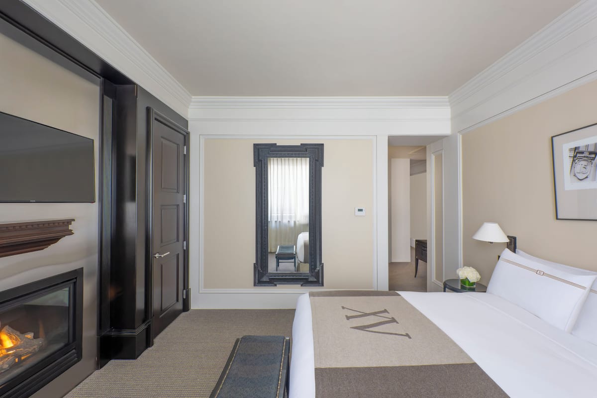 A hotel room with a bed, a fireplace, a mirror, and a nightstand with a lamp and flowers.