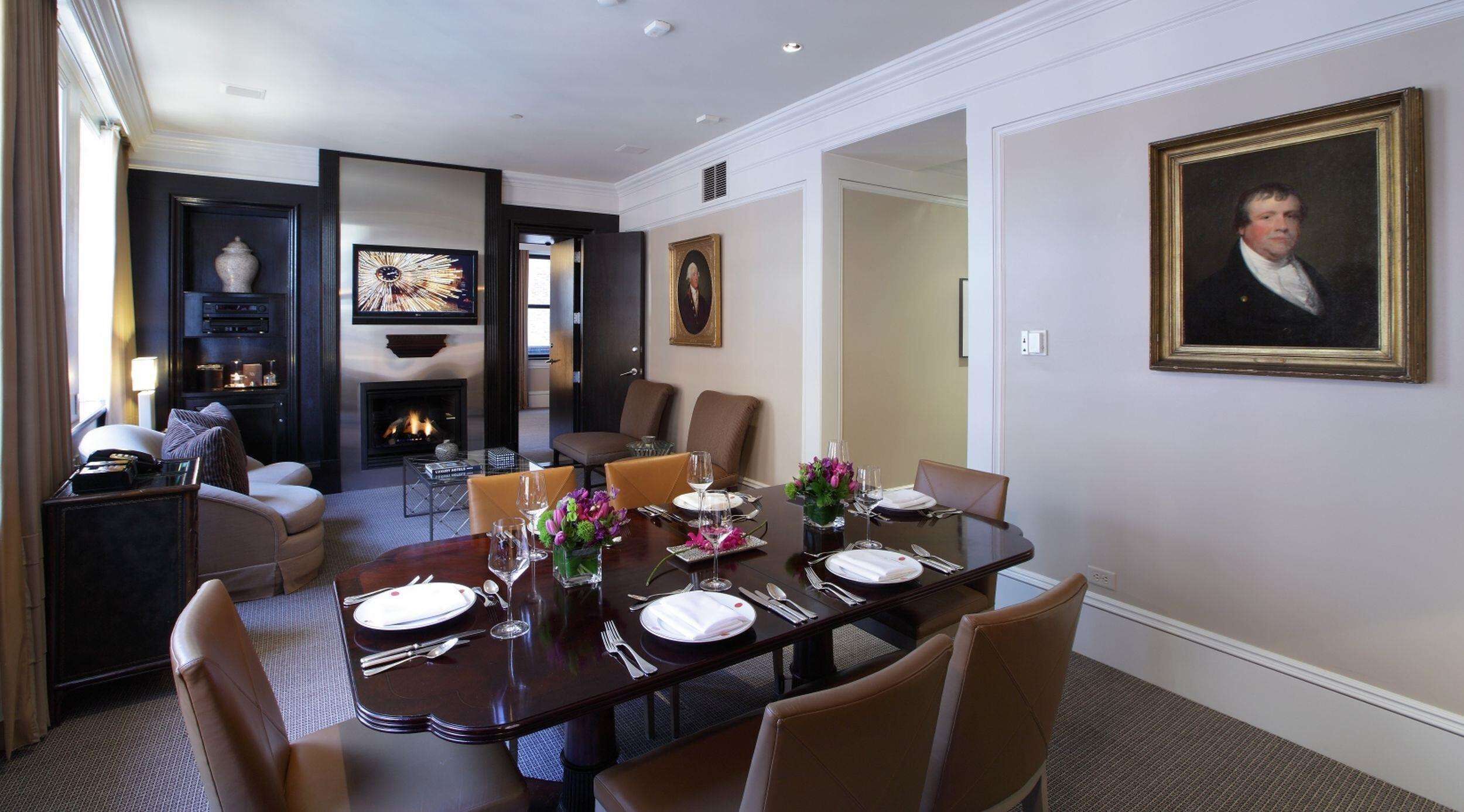 A dining room with a large table set for six, surrounded by chairs. There is a fireplace in the background, and a television mounted on the wall.
