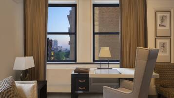 A hotel room with a desk, chair, lamp, and a view of the cityscape through large windows.