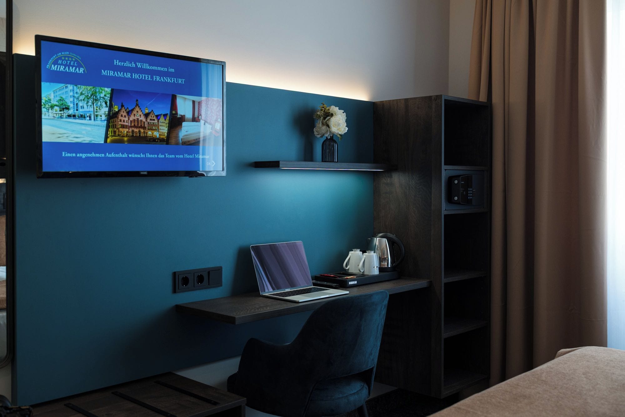 A hotel room with a TV displaying a welcome message, a desk with a laptop, a chair, and a shelf with a vase and a kettle.