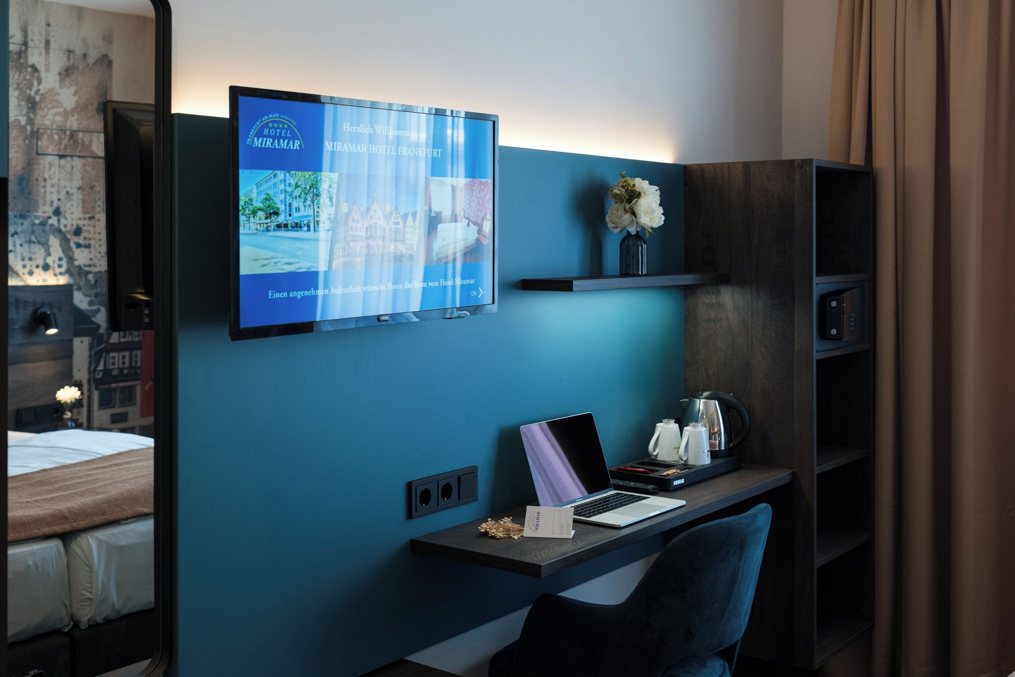A hotel room with a desk, laptop, and a television displaying the hotel's name.