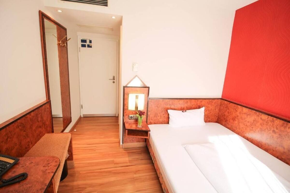 Habitación de hotel con piso de madera, cama con ropa blanca, mesita de noche con lámpara, espejo y pared de acento roja.