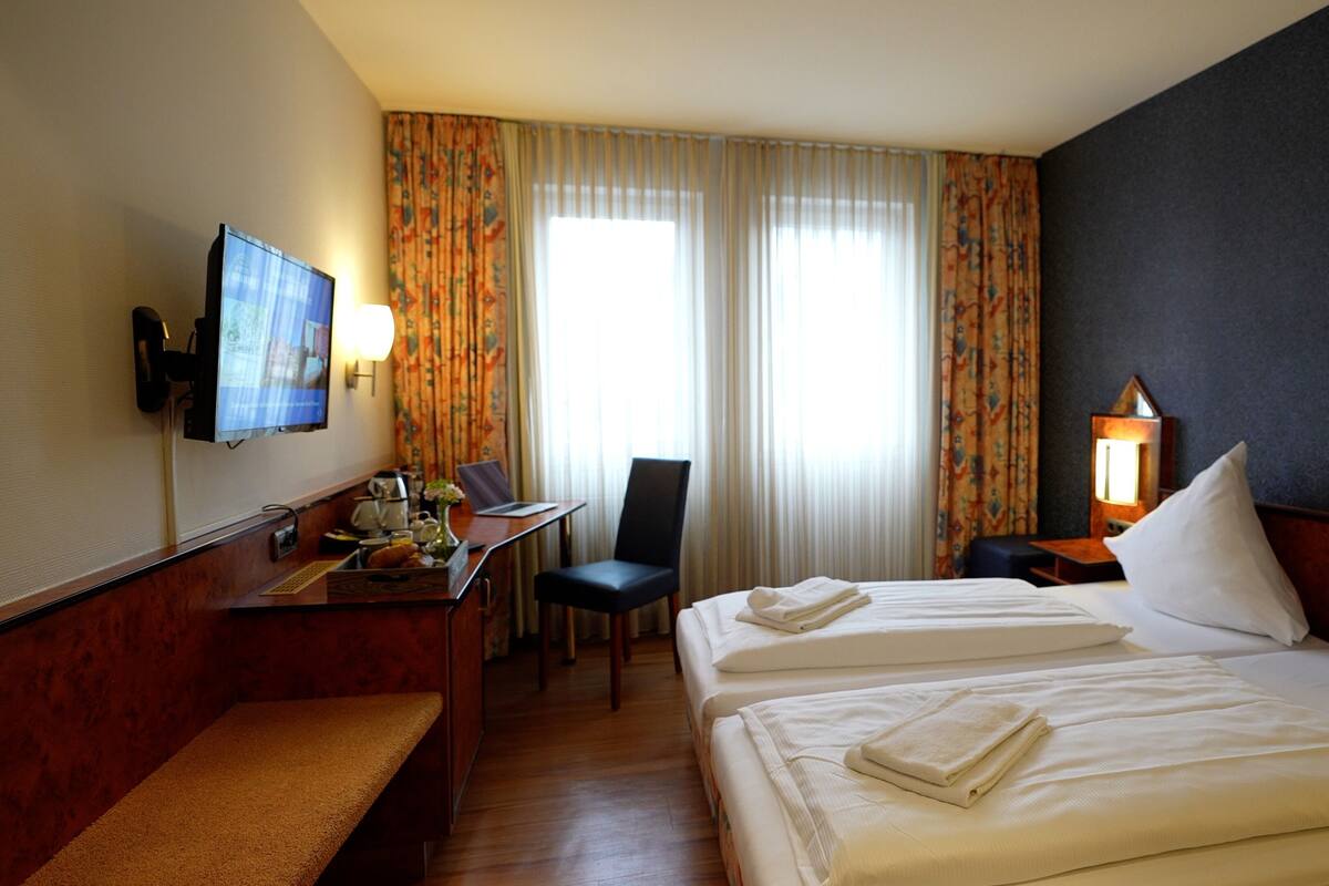 Habitación de hotel con dos camas, un escritorio con una computadora portátil, una silla, un televisor montado en la pared y una ventana con cortinas.