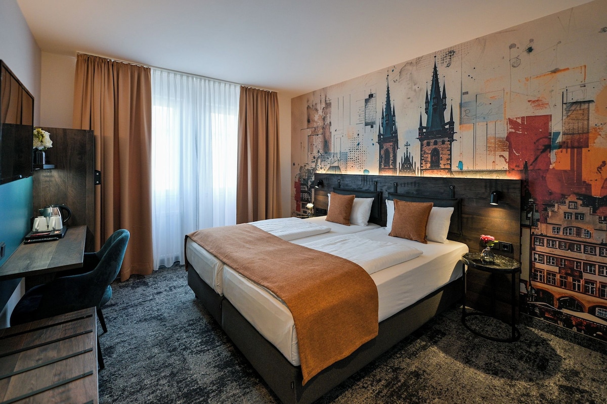 Una camera d'albergo con un letto grande, una scrivania con una sedia, un tavolino con un vaso e un murale raffigurante un panorama urbano.