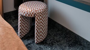Une petite pièce moderne, avec une étagère en bois, un tabouret à carreaux et une étagère murale ornée d’un vase de fleurs.