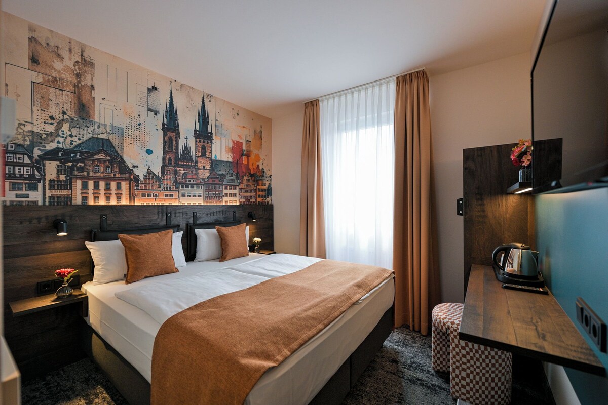 Una camera d'albergo con un grande murale raffigurante un panorama cittadino, un letto con coperta marrone, una scrivania con bollitore e una sedia.
