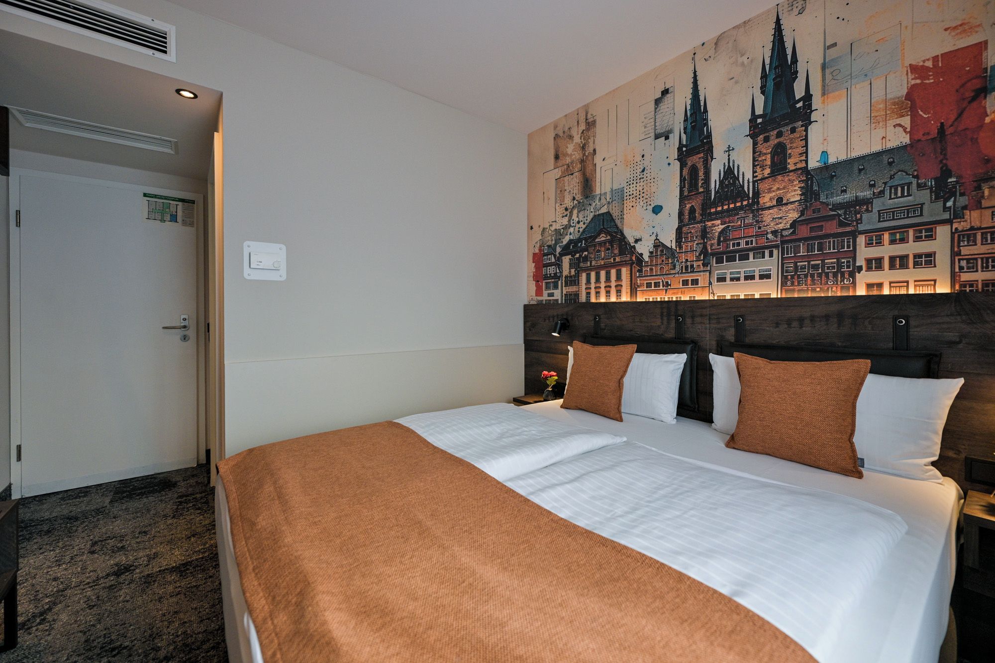 Habitación de hotel con una cama grande, cabecera con un mural de paisaje urbano, una puerta y una lámpara fijada a la pared.