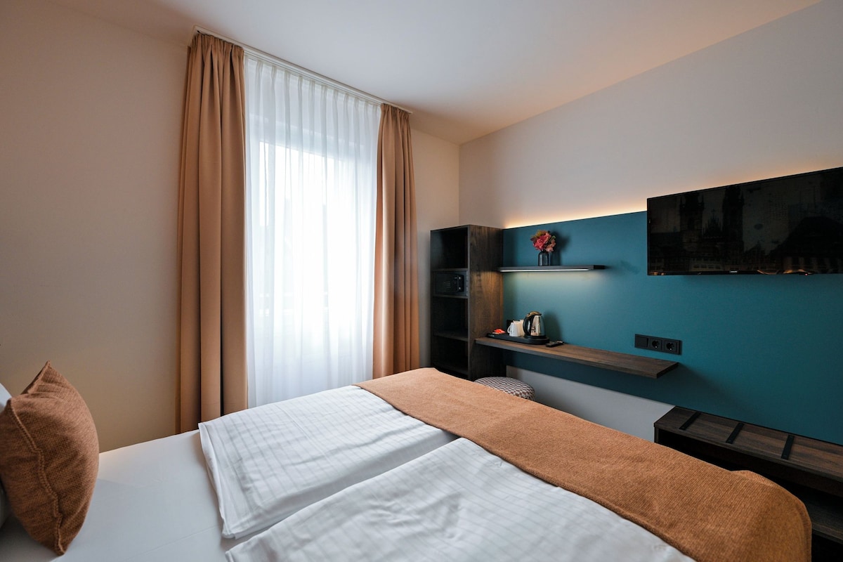 Una camera d'albergo con un letto, una TV fissata al muro, un piccolo ripiano con un vaso e una finestra con tende.