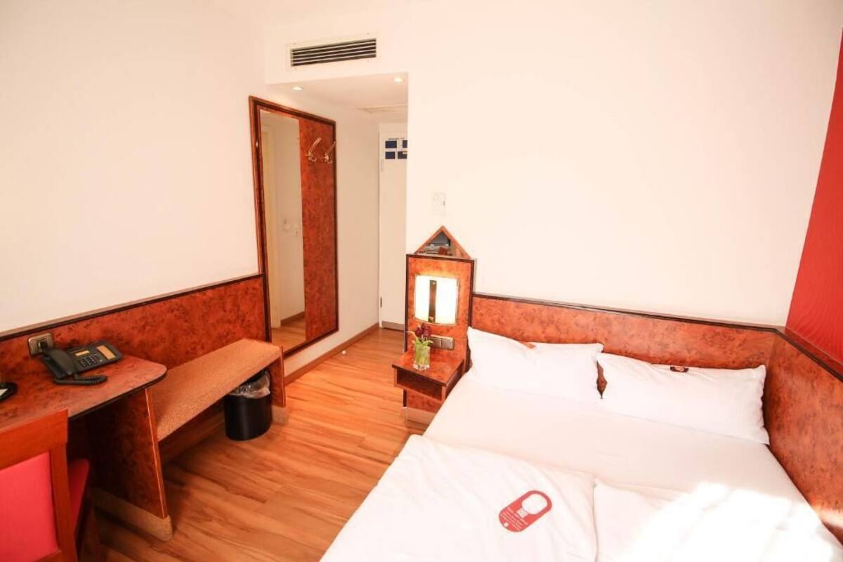 Habitación de hotel con piso de madera, una cama con ropa blanca, un escritorio con teléfono, una mesita con lámpara y un espejo en la pared.