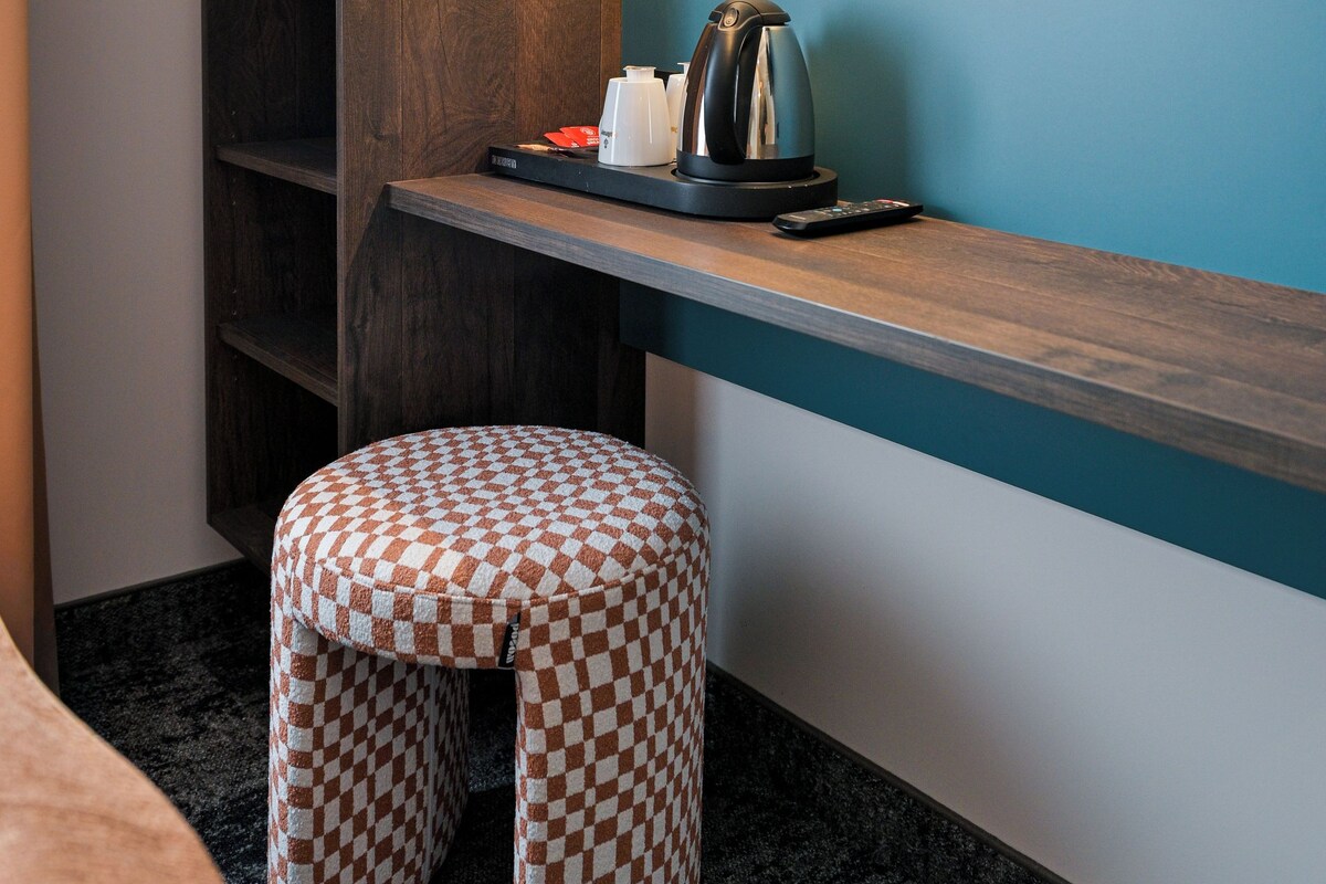 A compact hotel room with a wooden bookshelf, a small table with a kettle and remote control, a checkered ottoman, and a vase with flowers on a blue shelf.