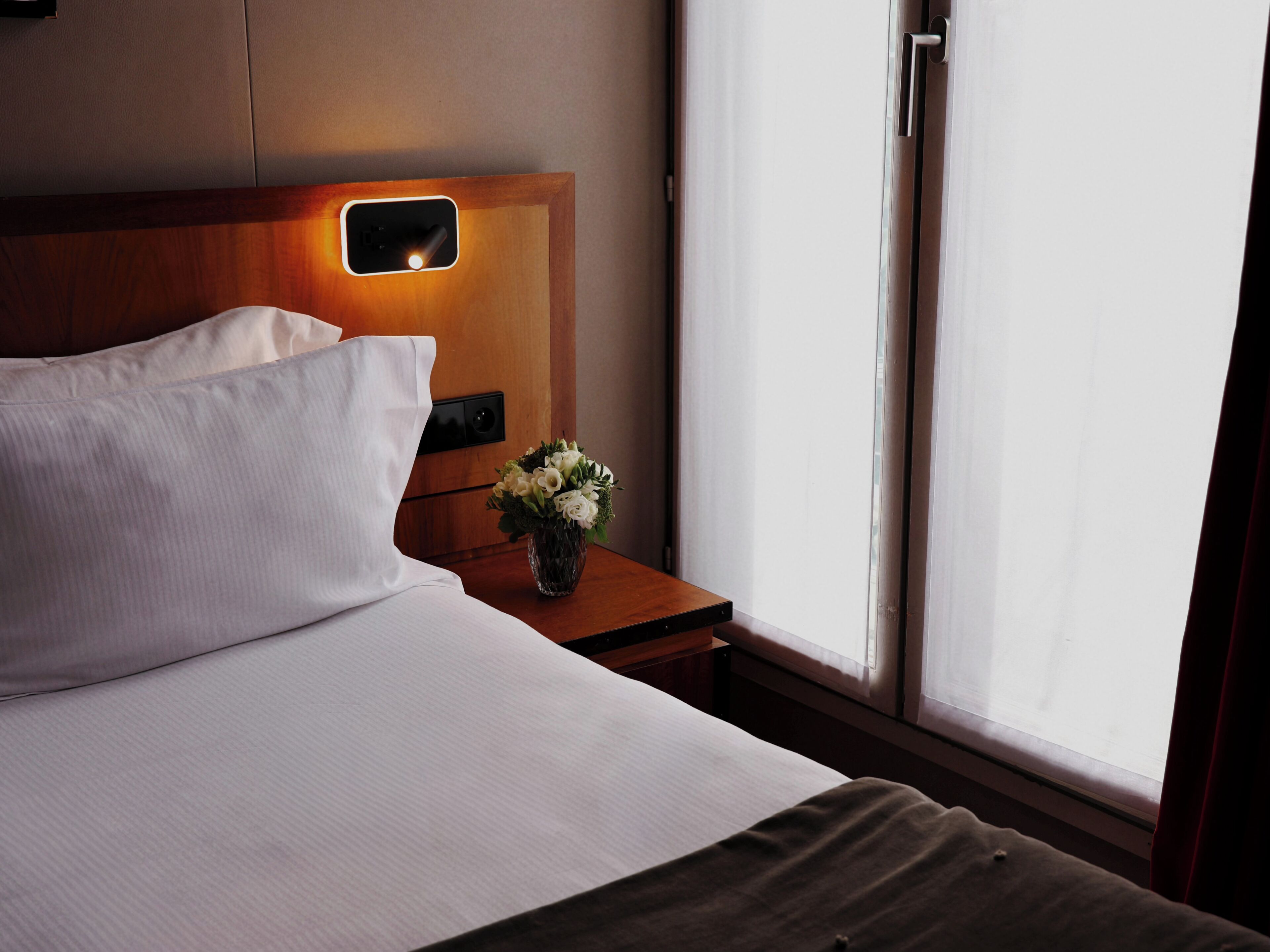 Een hotelkamer met een bed, nachtkastje, bloevas en raam met gordijnen.