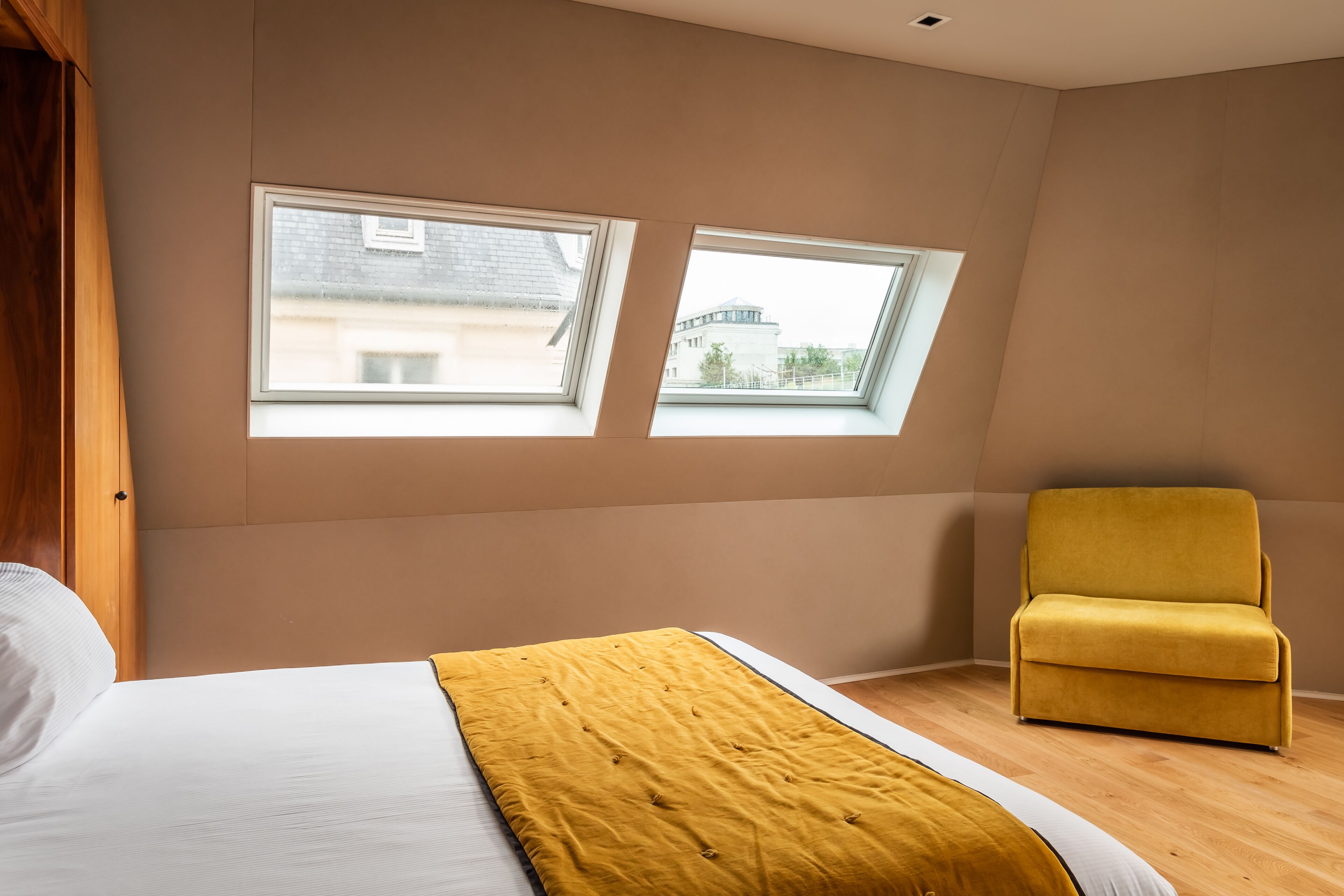 A bedroom with a bed, a yellow chair, and two windows with a view of buildings.